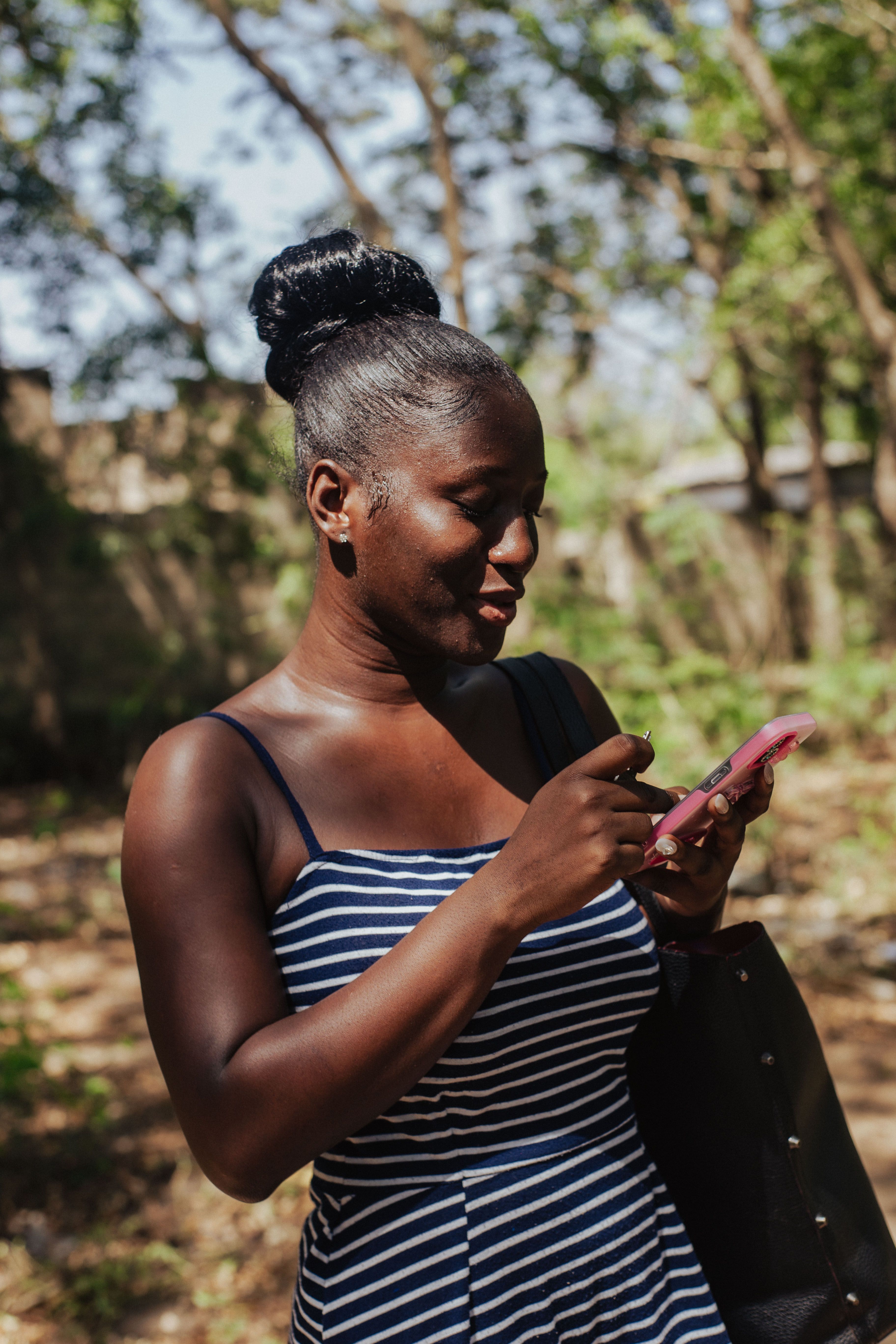 A woman in a striped dress using her phone while walking in a sunlit park in Accra, Ghana.