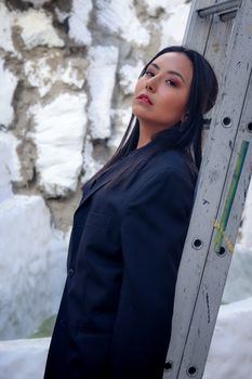 Fashionable woman in black coat leans against a ladder with a textured white stone background.
