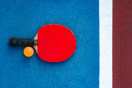 A red table tennis paddle and orange ball on a colorful court background.