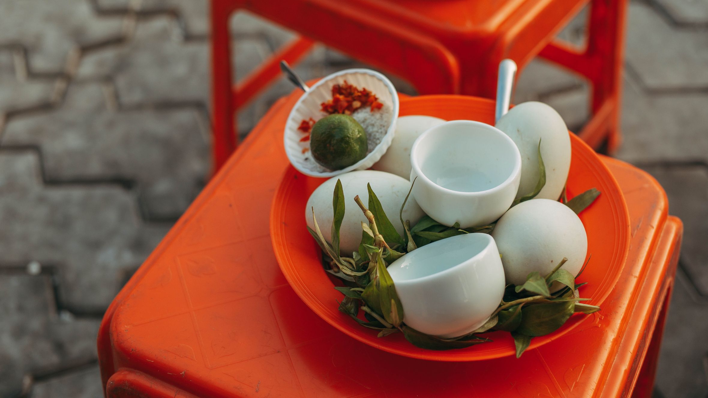 Close-up of a traditional street food setup featuring duck eggs, herbs, lime, and spices on an orange stool.
