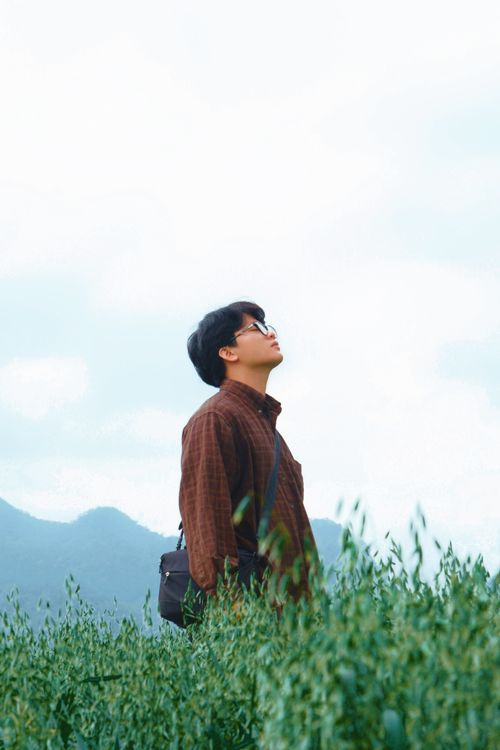 Free A man looks upwards while standing in a lush green field with mountains in the background. Stock Photo