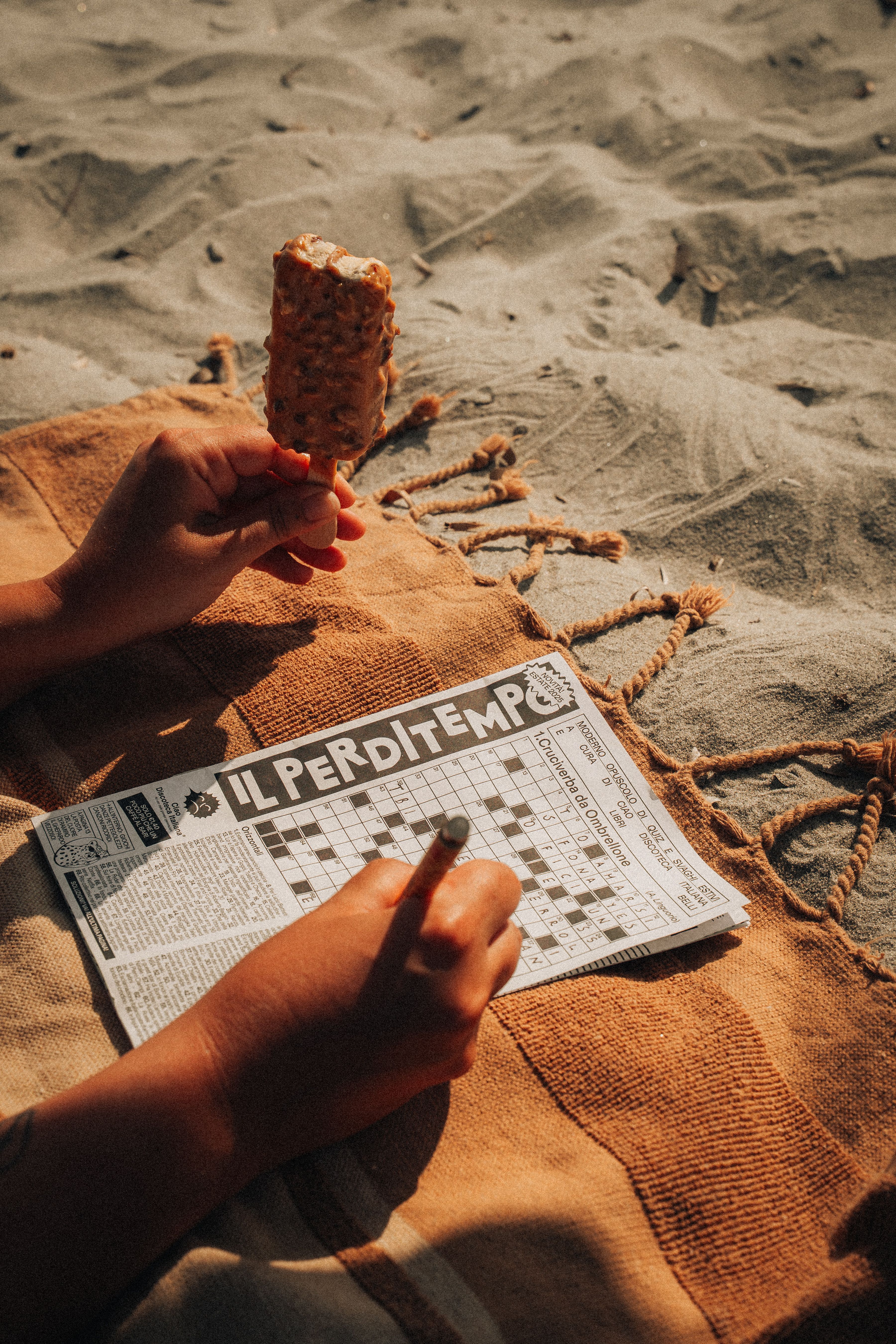 Person enjoying a crossword and ice cream on a sandy beach, with warm sunlight.