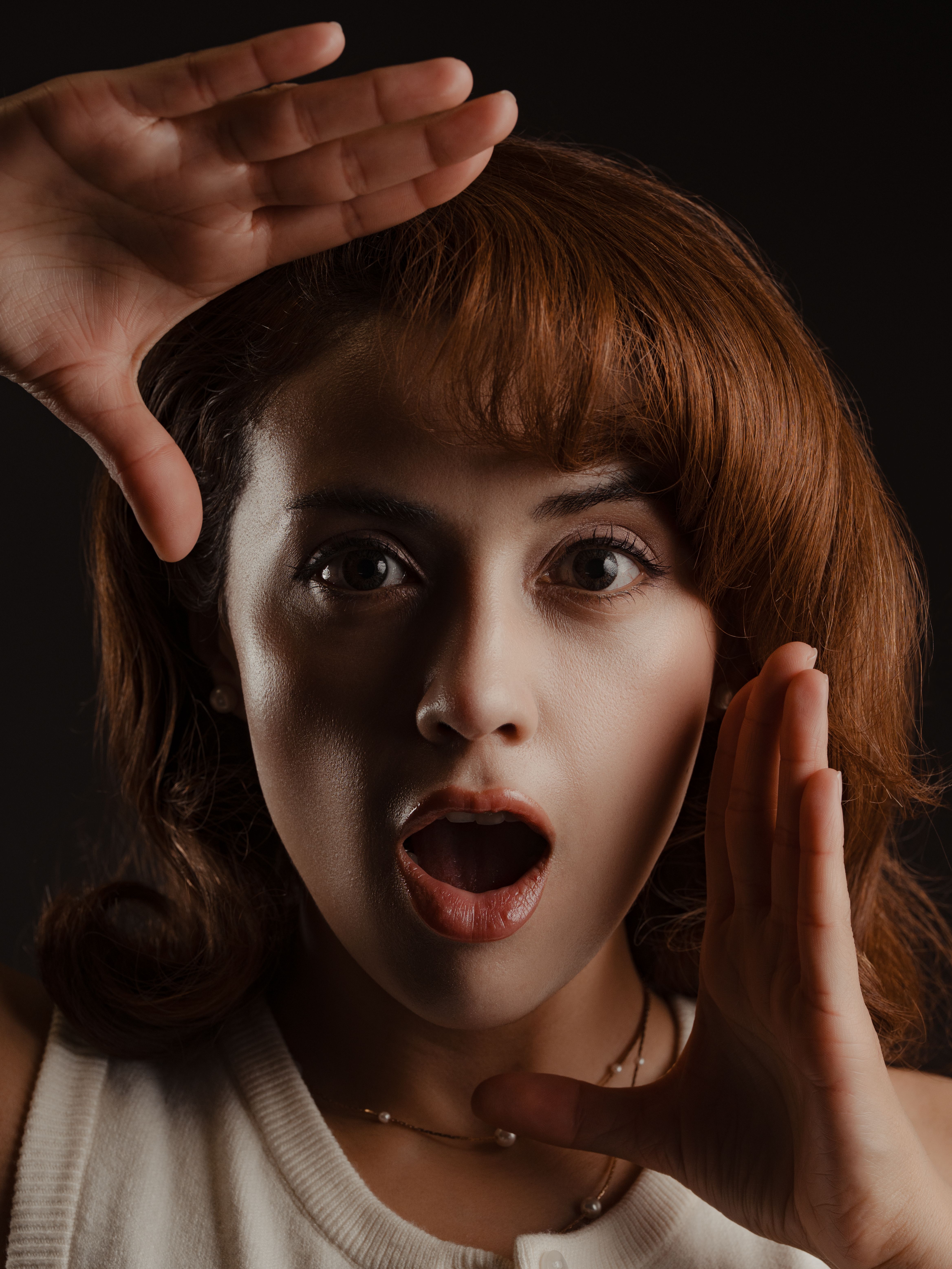Close-up portrait of an adult woman looking surprised with hands framing her face, in a dark setting.