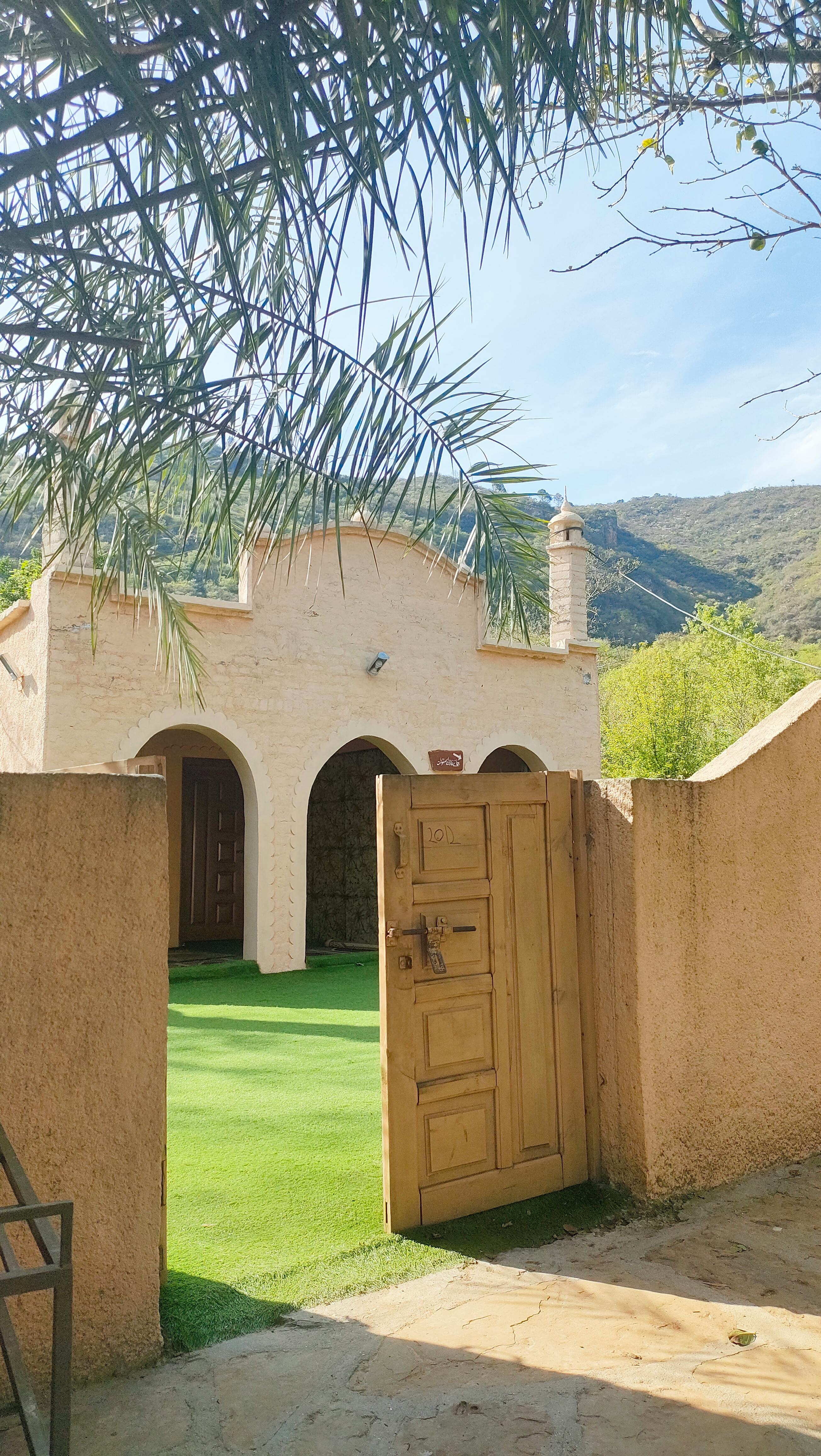 Rustic adobe home with open gate reveals lush garden under clear skies, framed by palm fronds.