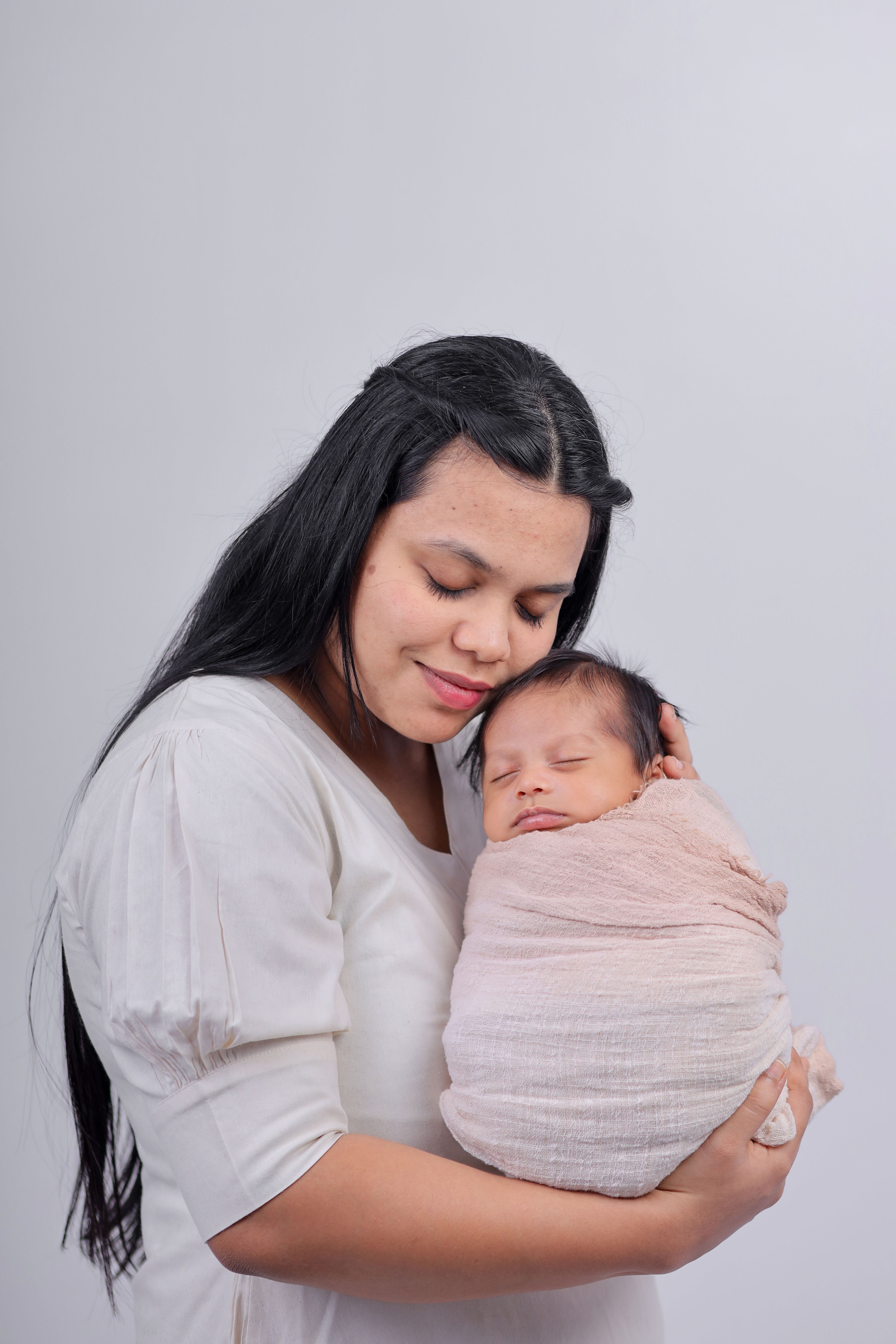 A heartfelt portrait of a mother lovingly holding her wrapped newborn baby against a neutral backdrop.