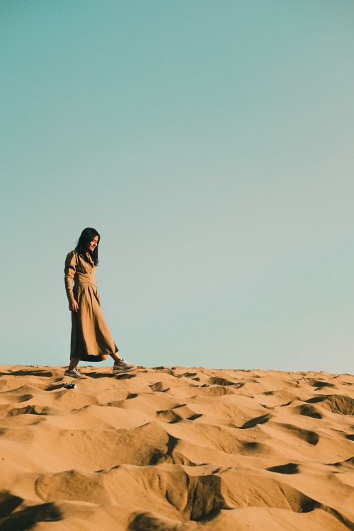 Free A woman walks across vast golden sand dunes under a clear blue sky, capturing serene desert beauty. Stock Photo