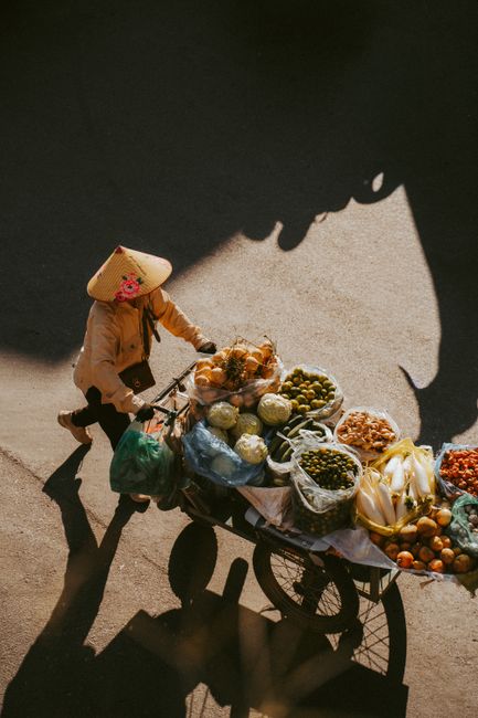 Street vendor pushing a bicycle cart loaded with various fruits and vegetables under warm sunlight in Hanoi, Vietnam.