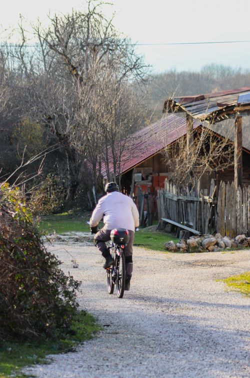 Free A cyclist enjoys a leisurely ride on a gravel path through a rural area with rustic buildings. Stock Photo