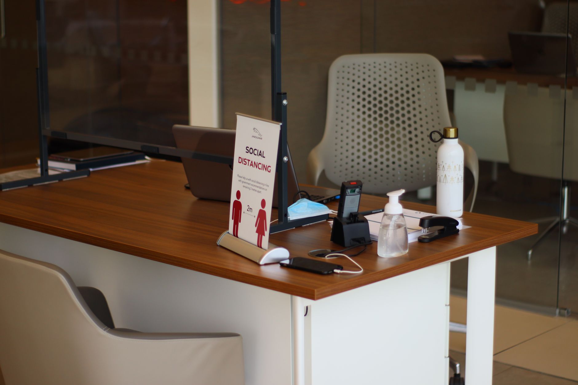 Office desk featuring laptop, sanitizer, and social distancing sign during the day.