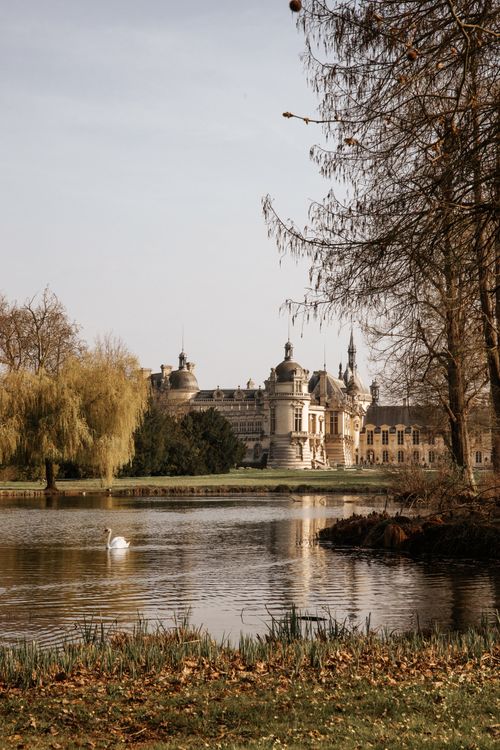 Free Château de Chantilly reflected in a tranquil lake with a white swan swimming amidst autumn scenery. Stock Photo