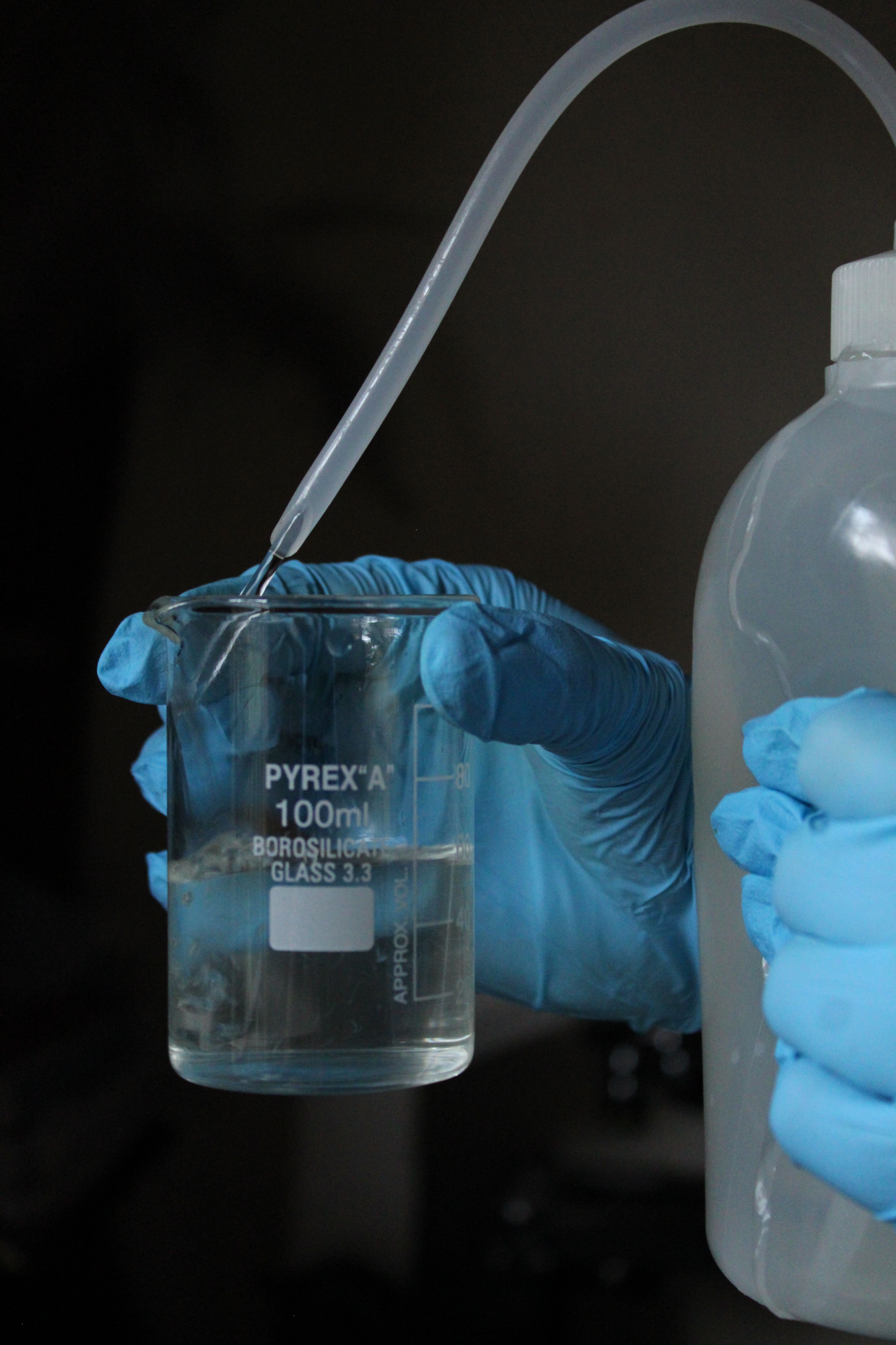 Blue gloved hands pouring liquid into a 100ml borosilicate glass beaker using a plastic bottle.
