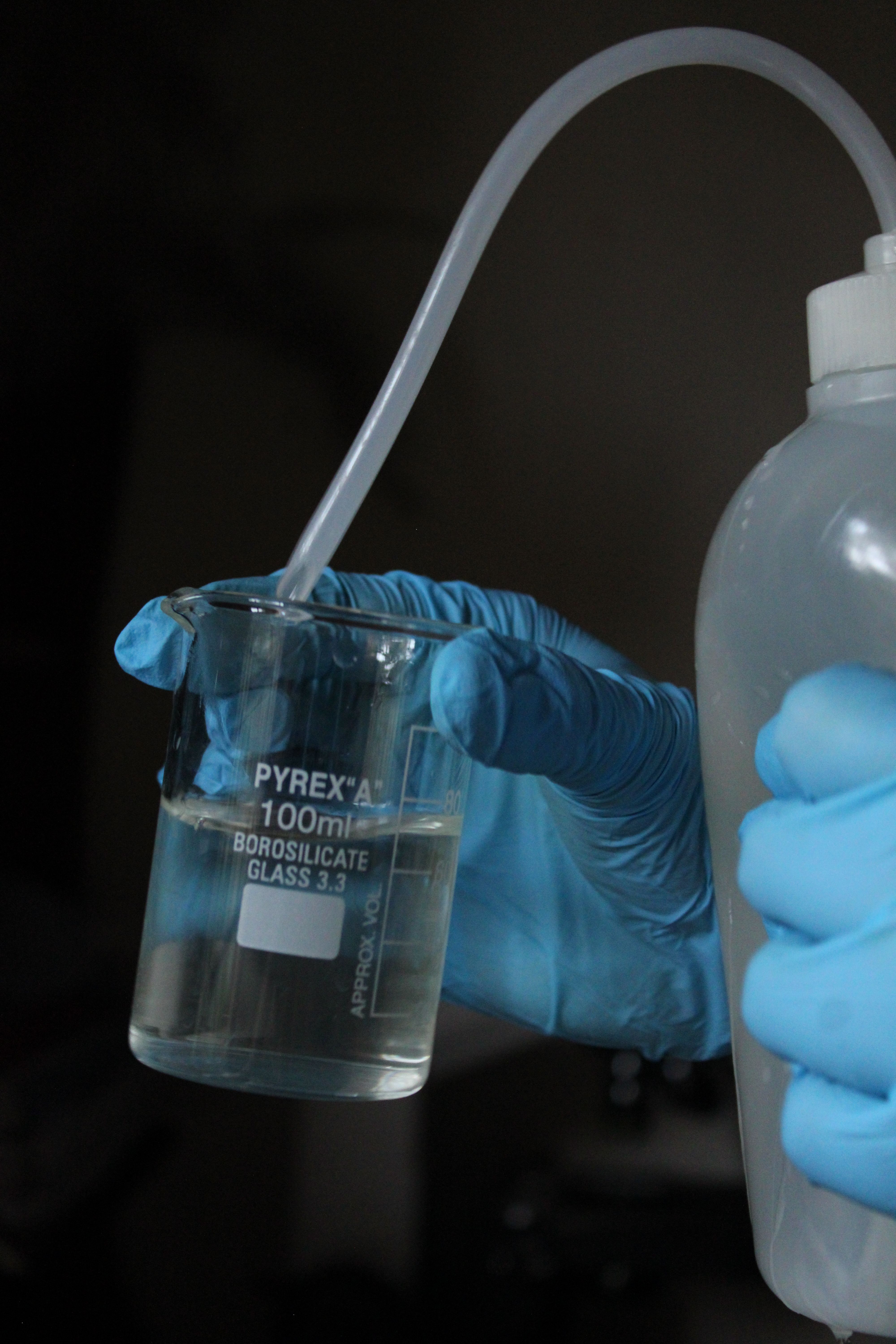 Close-up of gloved hands pouring liquid into a glass beaker in a lab setting.