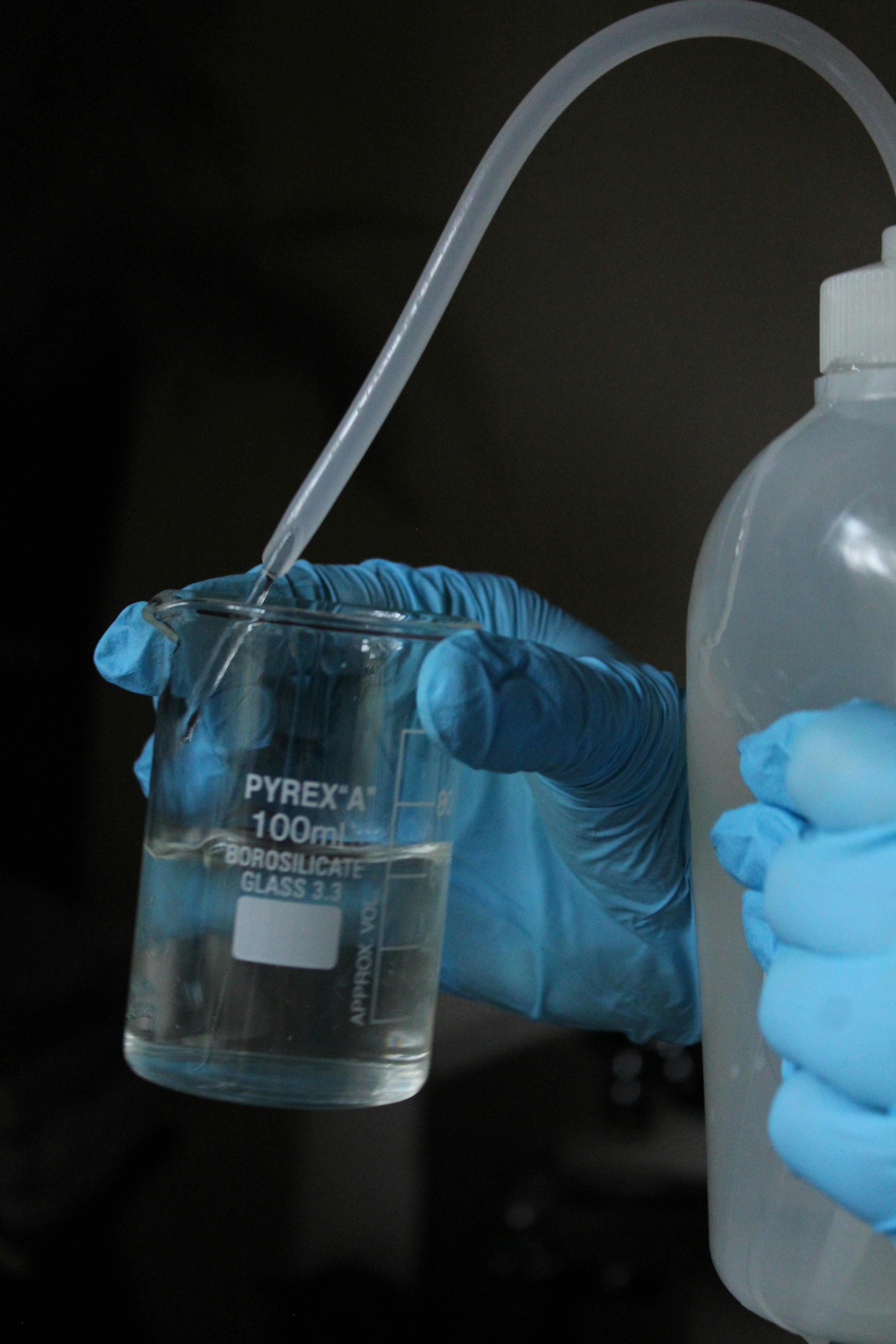 Scientist in gloves pouring clear liquid into a 100ml Pyrex beaker for an experiment.