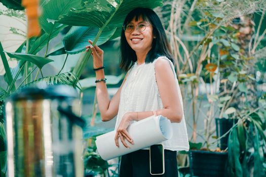 Young woman smiling among lush green plants, holding a yoga mat outdoors.