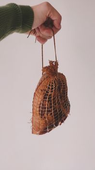 Hand holding a cured ham wrapped in netting against a neutral background.