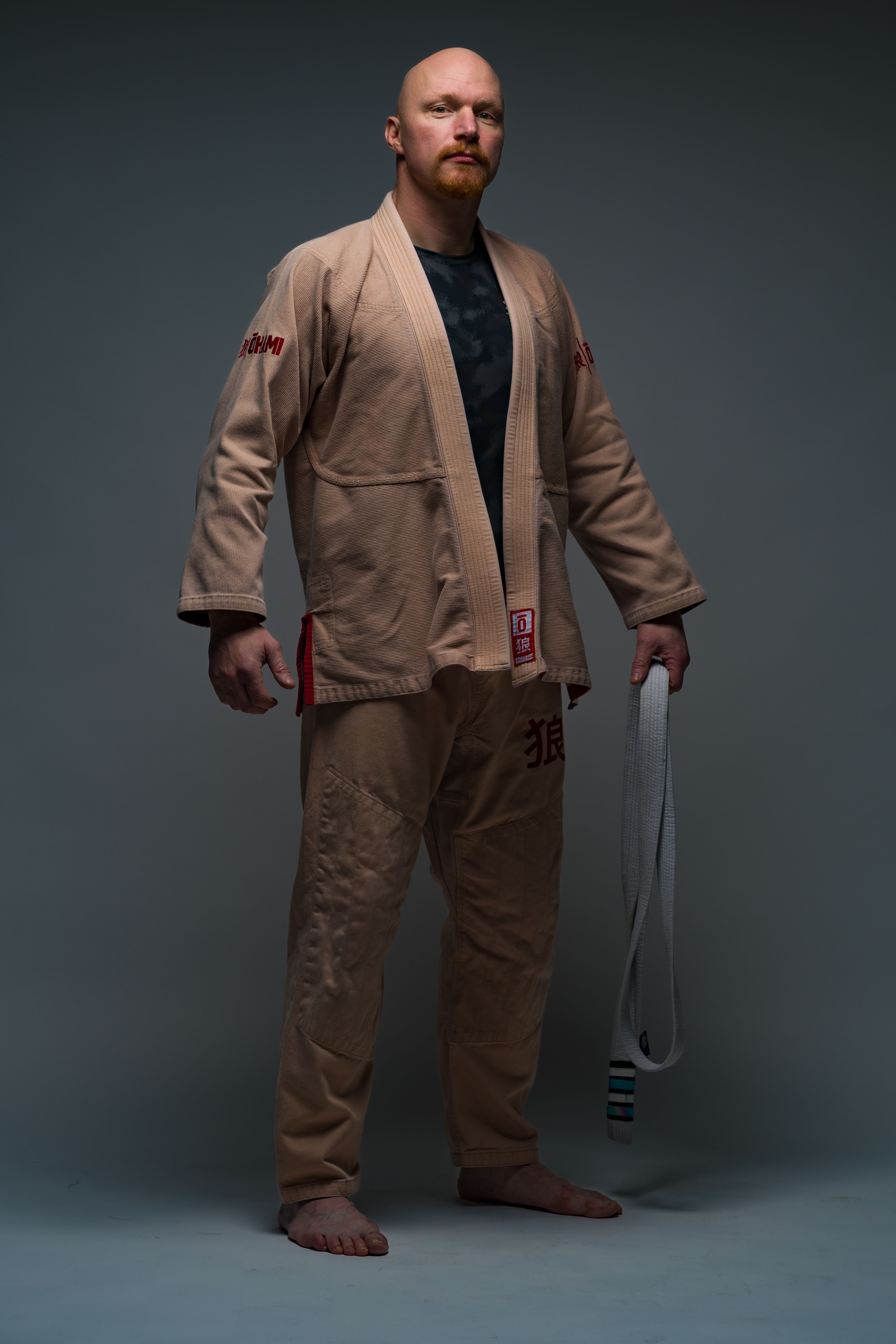Portrait of a martial artist in a beige gi holding a white belt, posing confidently indoors.