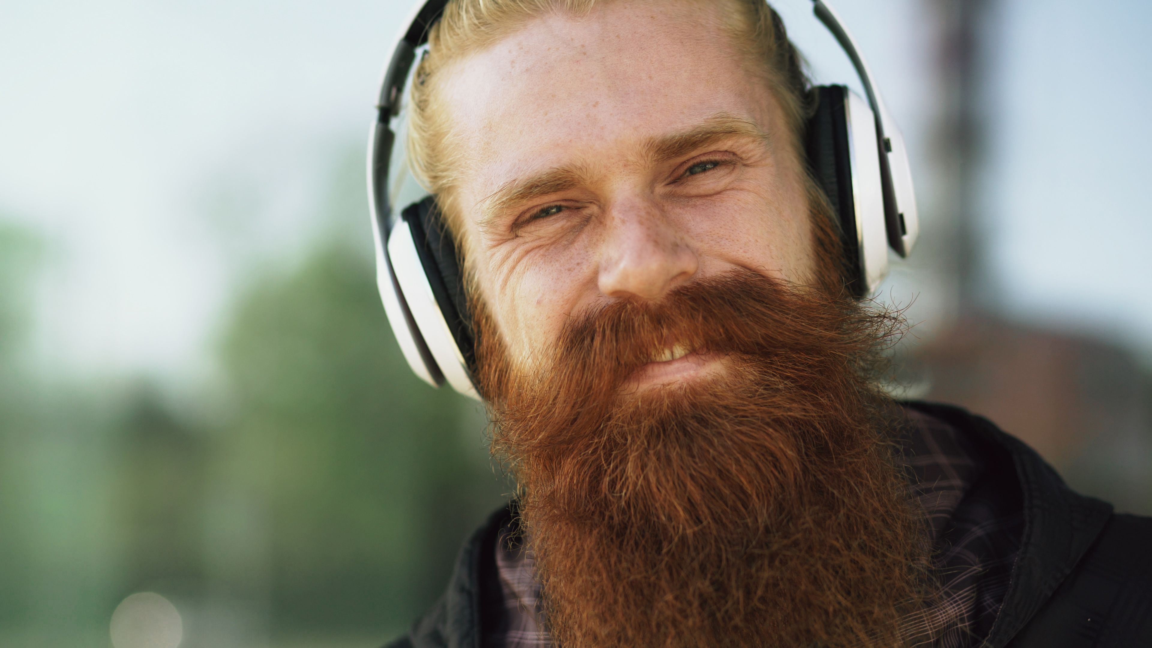 Close-up of a smiling man with a red beard wearing headphones, enjoying music outdoors.
