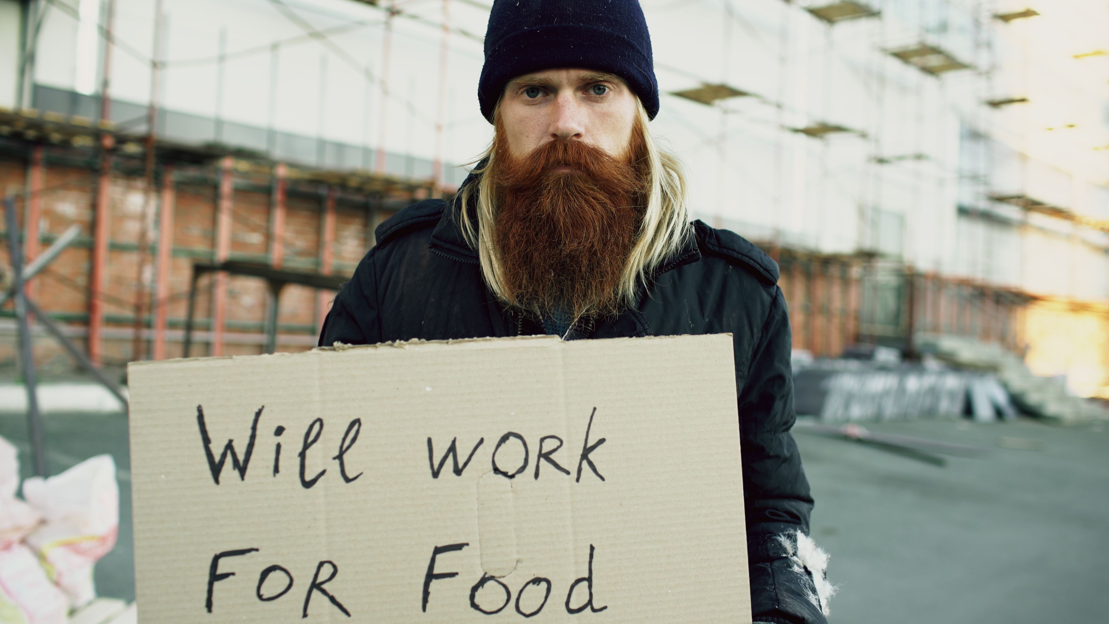A man with a long beard holds a 'Will Work for Food' sign outdoors with construction scaffolding in the background.