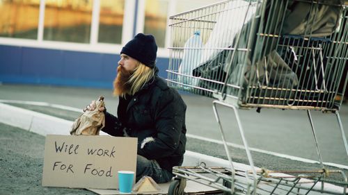 Free A homeless man sits outdoors with a sign seeking work for food. Stock Photo