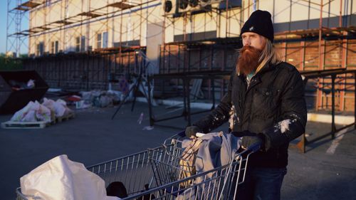 Free Bearded man pushes shopping cart near construction area in evening light. Stock Photo