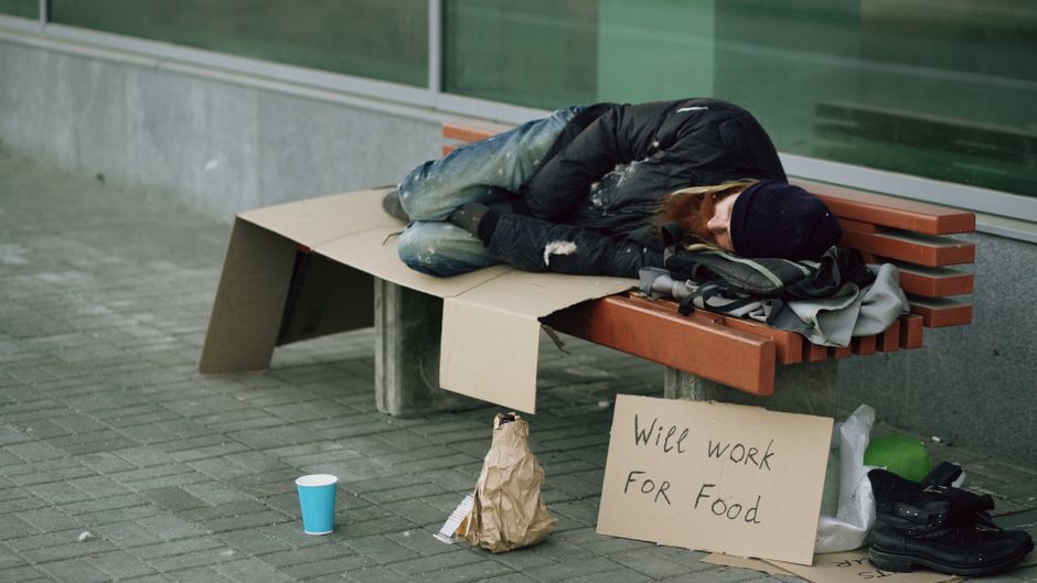 A homeless person sleeps on a bench with a sign reading 'Will work for food'.