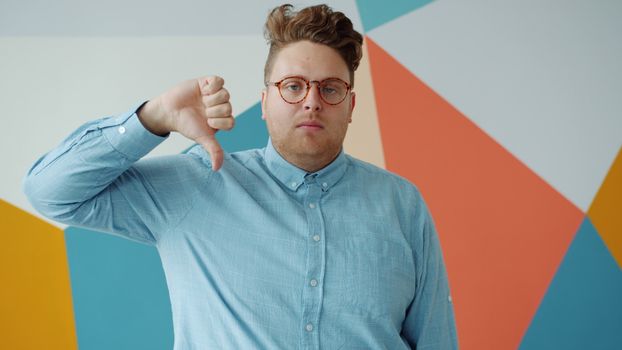 Young man in blue shirt gives a thumbs-down against colorful geometric background.