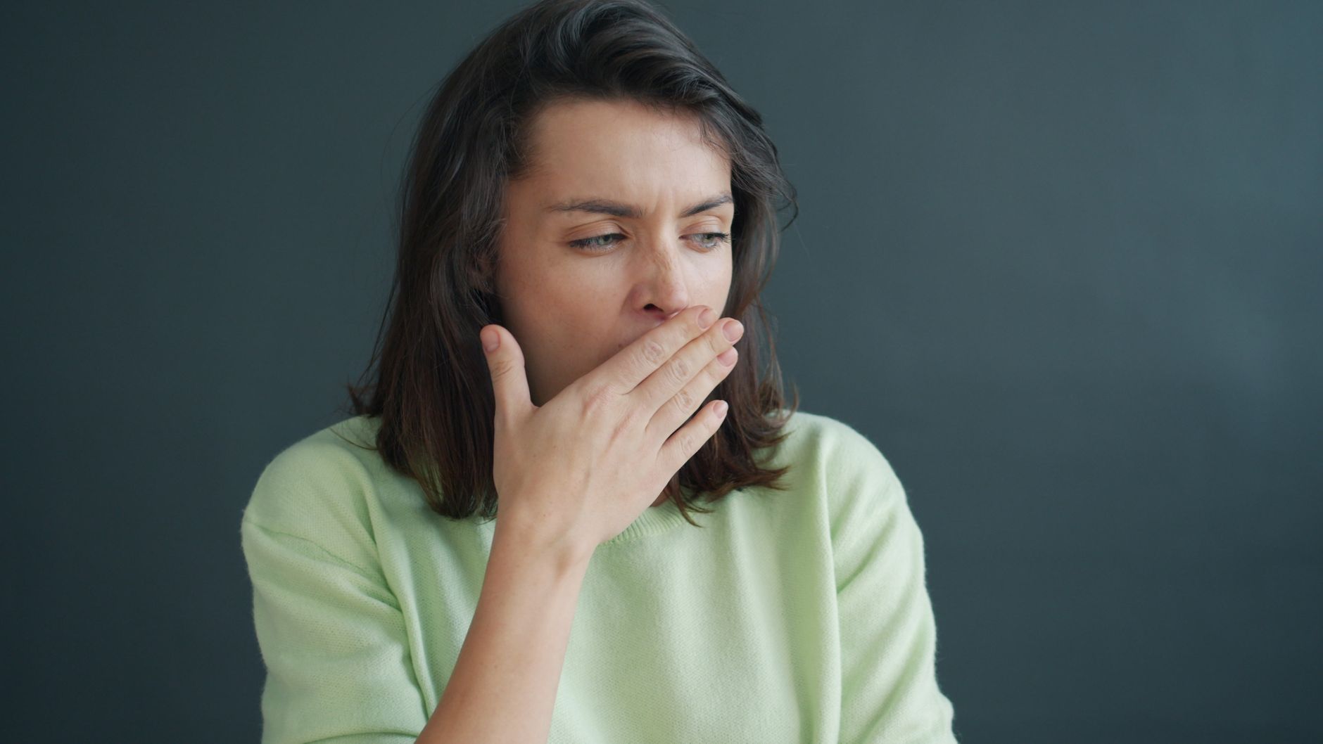 Young woman yawning, covering mouth with hand, wearing casual sweater against a gray background indoors.