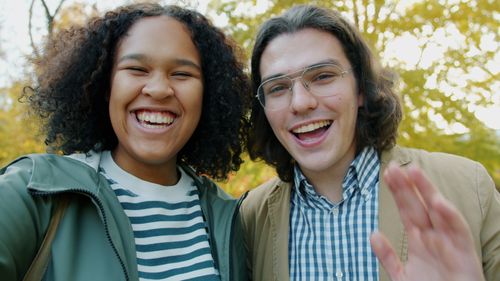 Free Two friends happily smiling and waving in a vibrant autumn park. Stock Photo