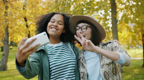 Free Young friends smiling and taking selfies in a vibrant autumn park. Stock Photo