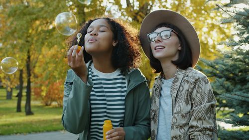 Free Two friends enjoy a sunny fall day blowing bubbles in a park, surrounded by vibrant autumn foliage. Stock Photo