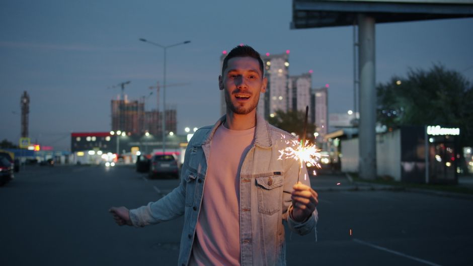 A young man holding a sparkler in an urban setting at dusk, celebrating.