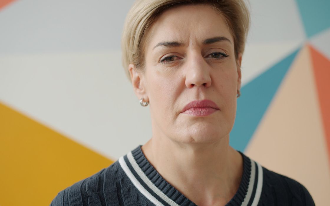 Middle-aged woman with a serious expression, forehead and brows clearly visible, in front of a colourful wall