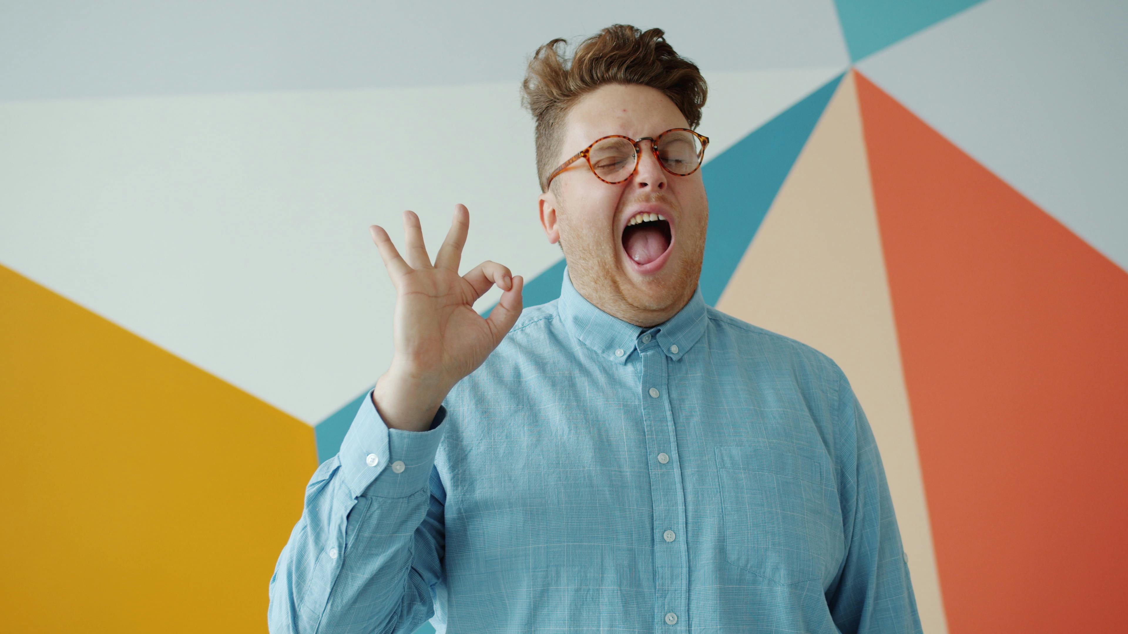 Smiling man in blue shirt making an okay sign against colorful wall.