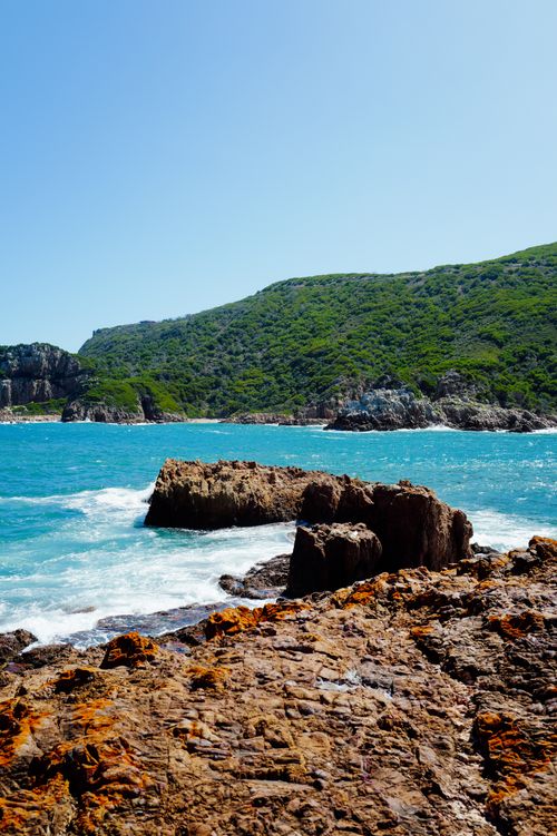 Free Vibrant coastal scene featuring rocky shoreline and lush hills under a bright blue sky. Stock Photo
