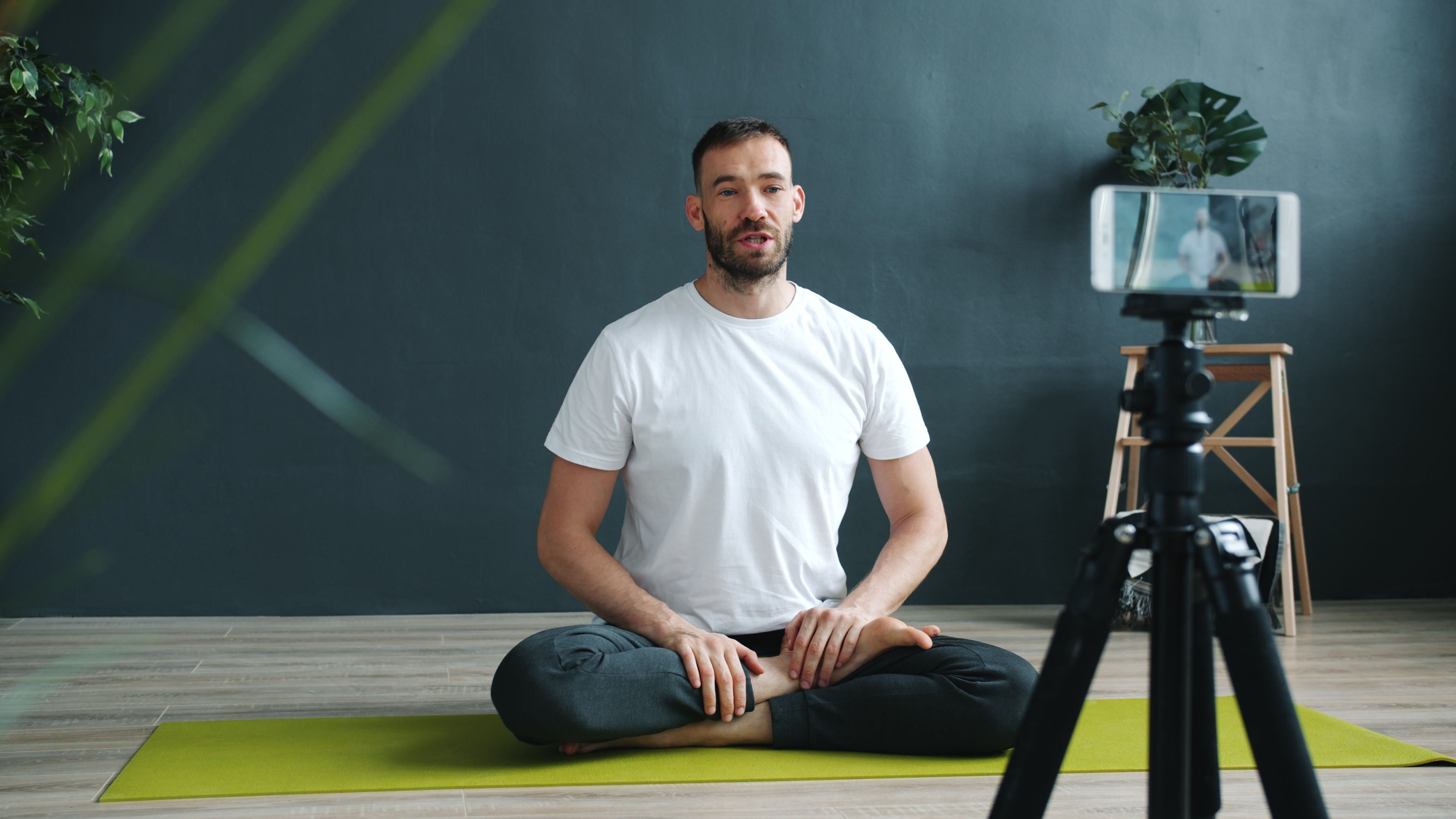 A man in white T-shirt filming an online yoga session indoors using a smartphone on a tripod.