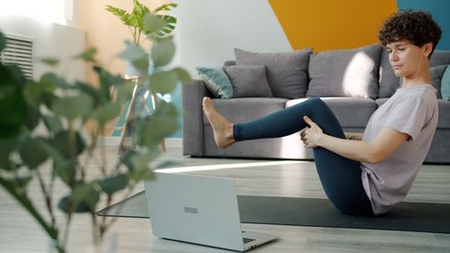 Free Woman doing yoga at home with focus on laptop. Modern room setting for virtual workout. Stock Photo