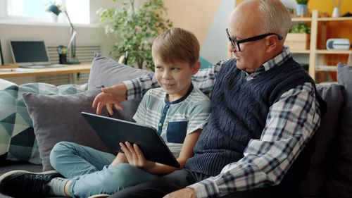 Free A senior adult and child bonding over a tablet while sitting on a couch indoors. Stock Photo