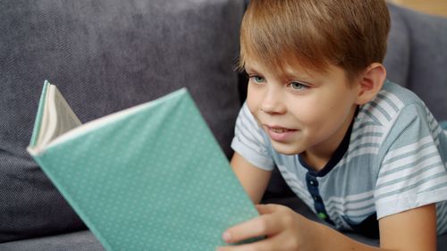 Free A child enjoys reading a teal book while relaxing on a sofa, focused on the story. Stock Photo