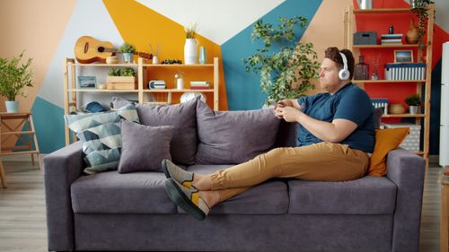 Free Young man with headphones gaming on a cozy grey sofa in a modern living room. Stock Photo