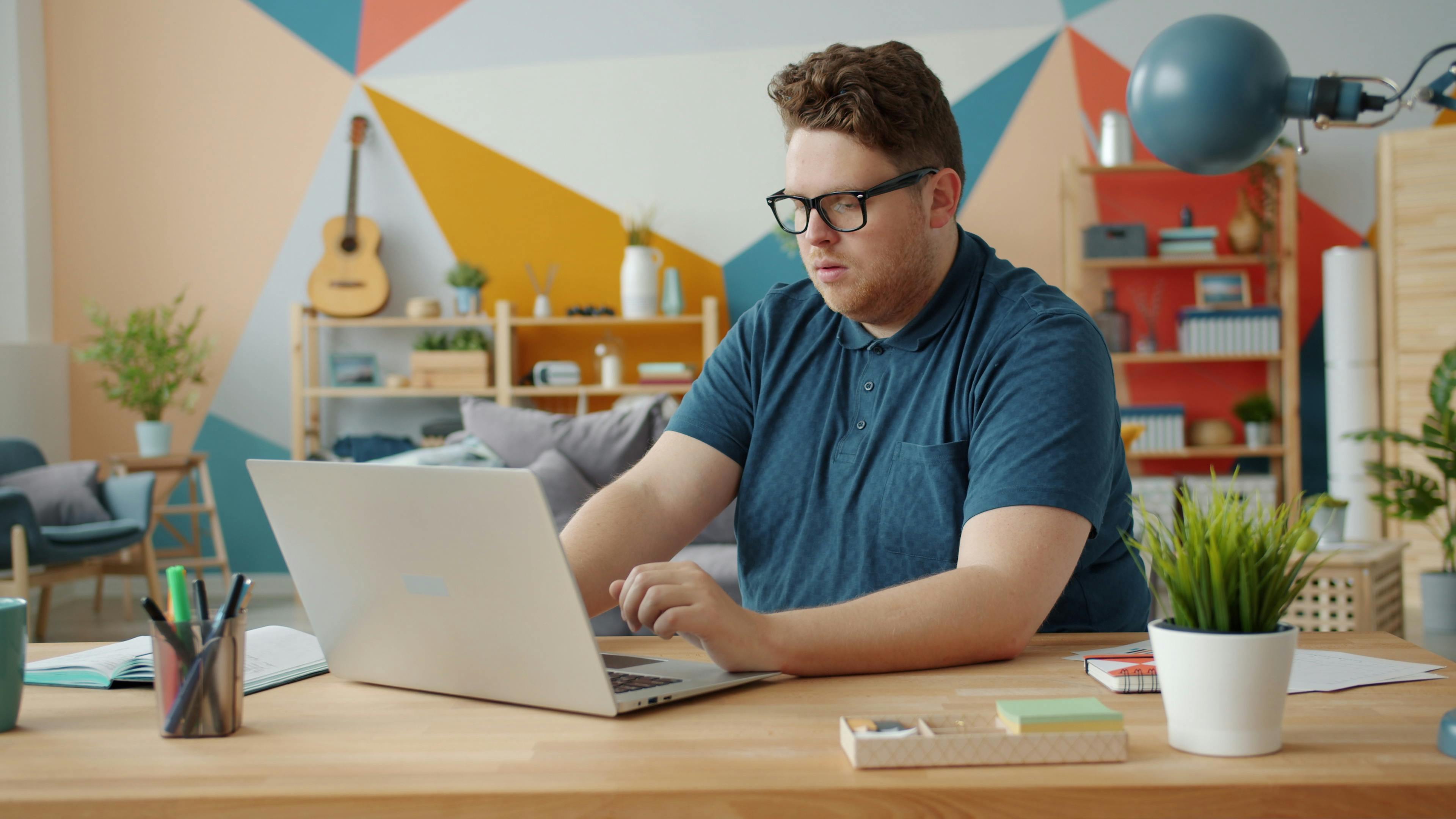 Young bearded man focused on work at home office desk with laptop.