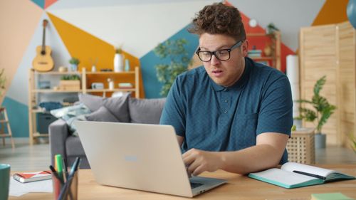 Free Focused man using a laptop in a colorful, modern home office environment, surrounded by artistic decor. Stock Photo