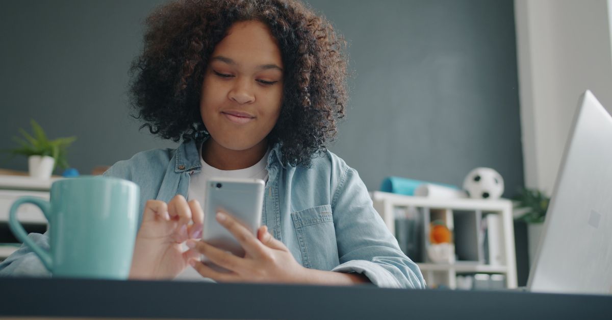 Gen Z person checking phone with laptop showing passive income dashboard, modern apartment background