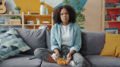 Free Young woman sitting on couch with snacks in a modern living room. Stock Photo