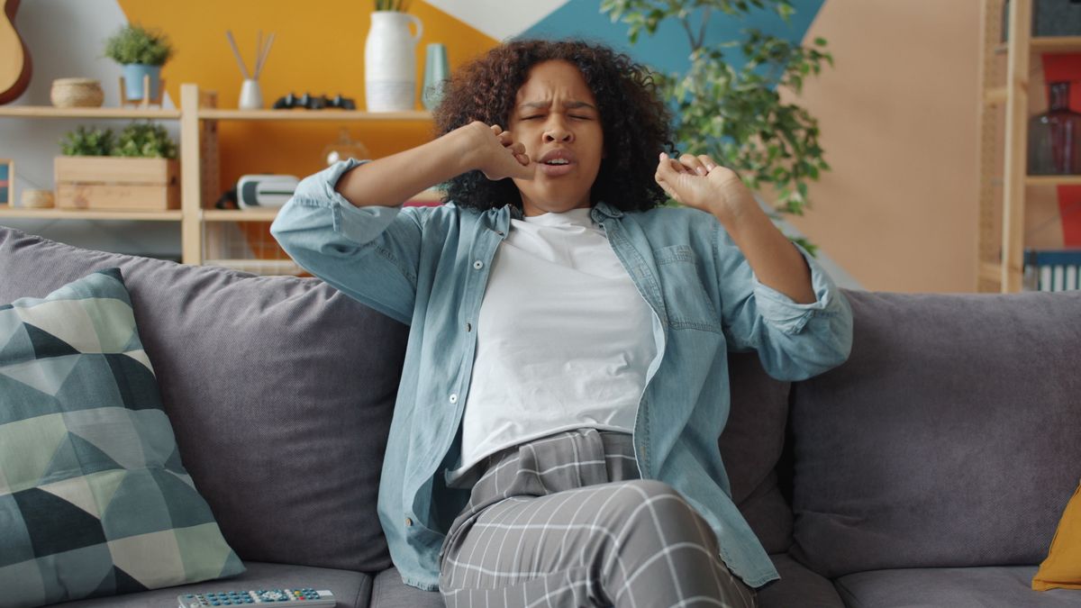 A woman yawning on a living room sofa, wearing comfortable casual clothes