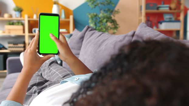 Woman lying on couch holding a green screen smartphone indoors, ideal for apps.