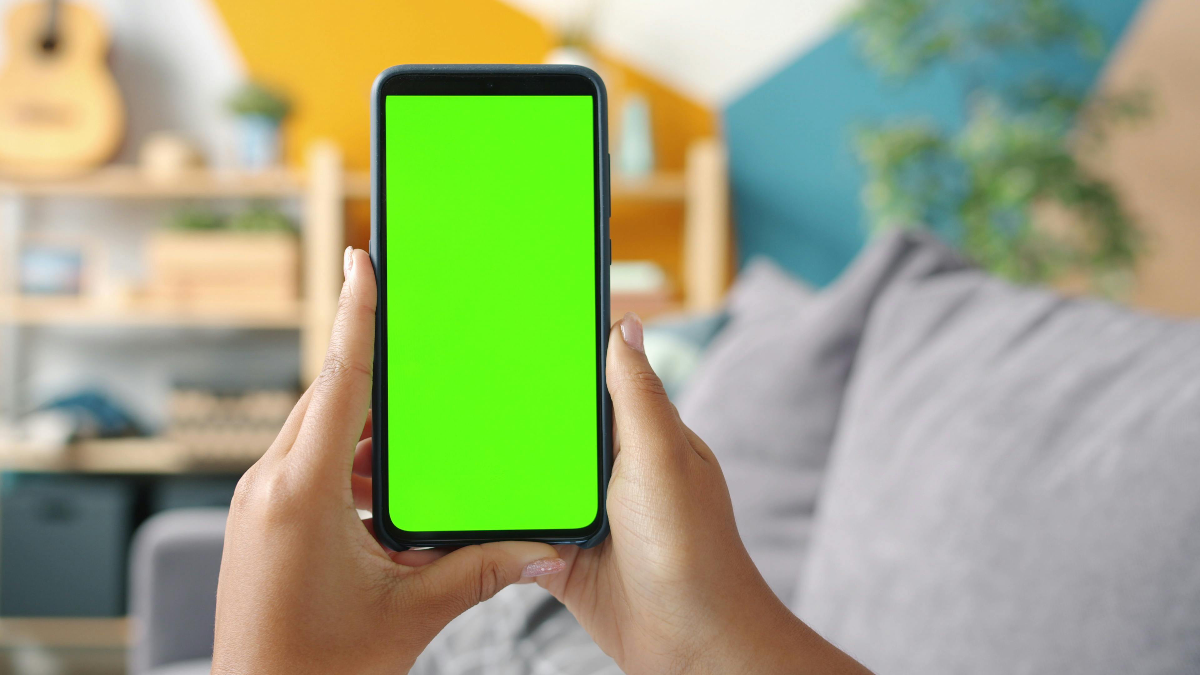 Hands holding smartphone with green screen in modern living room.