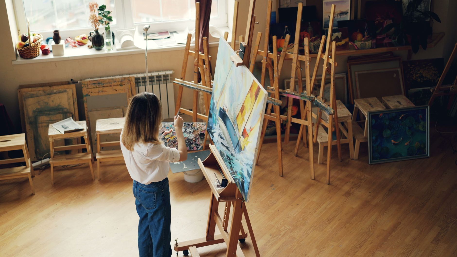 A woman artist paints on a canvas in a bright, well-equipped studio.