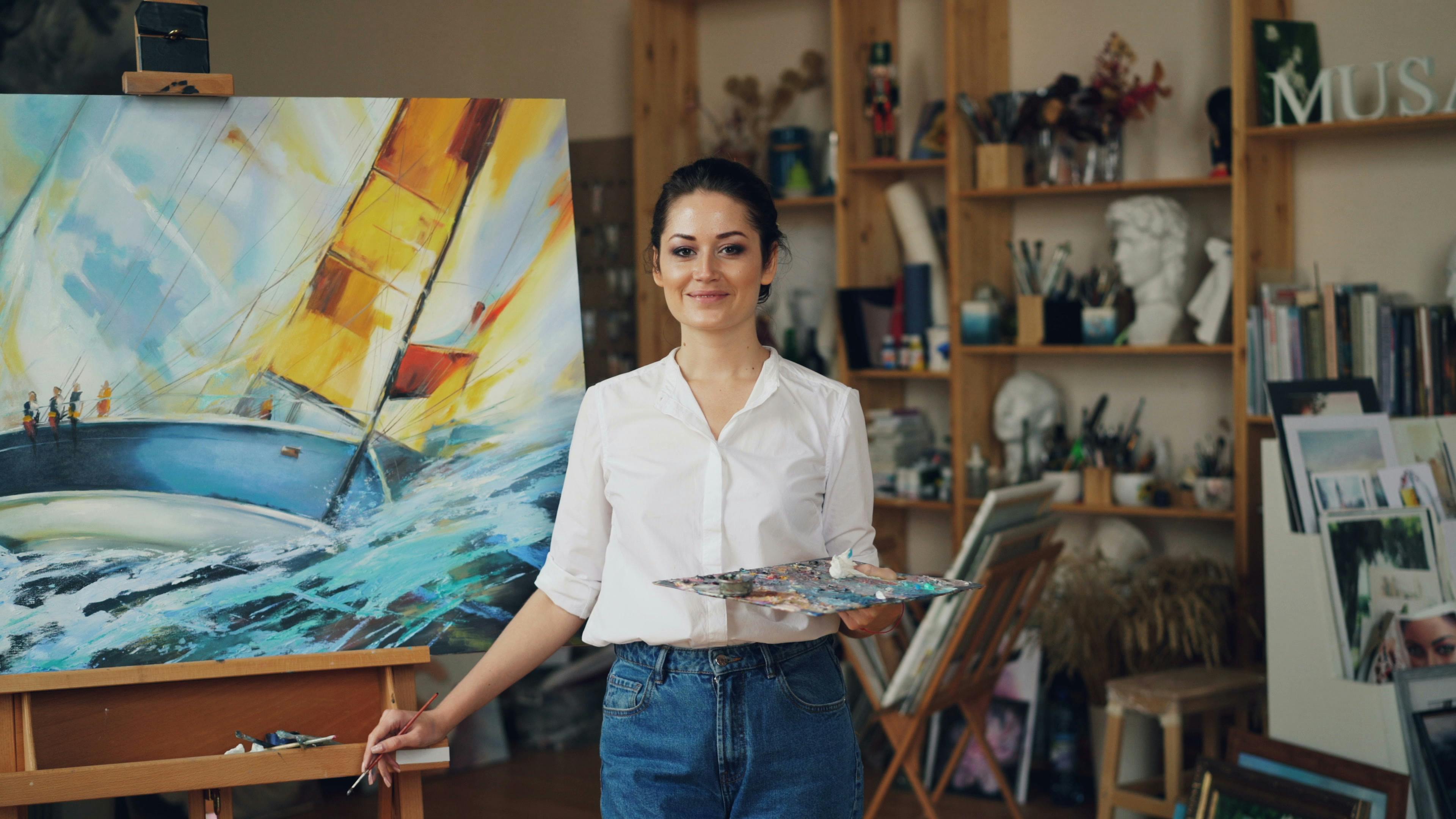 Female artist in studio smiling in front of her vibrant sailboat painting, holding a palette.