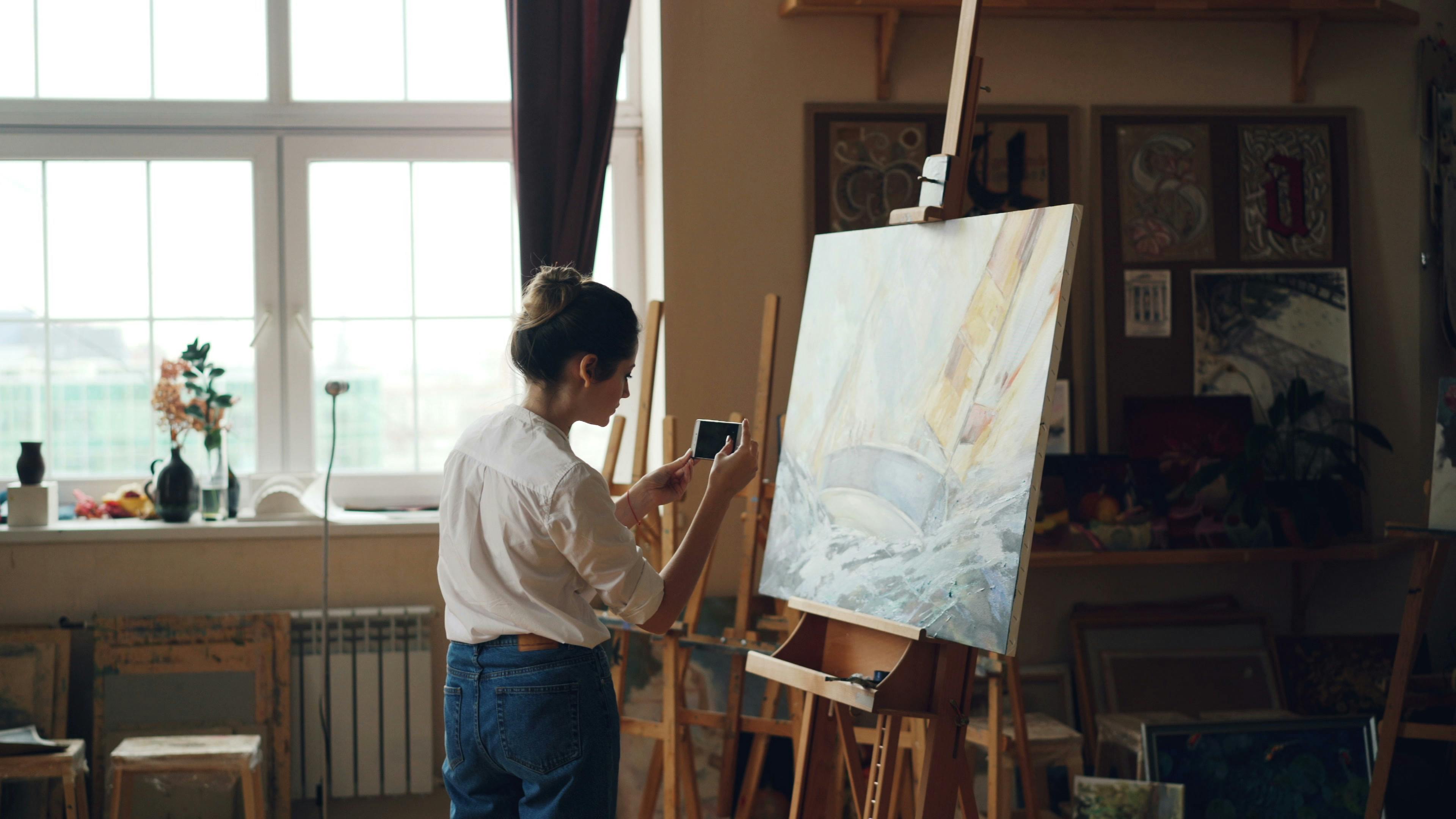 A person capturing an artistic painting of a sailboat in a well-lit art studio.