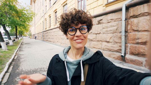 Smiling woman with glasses taking a selfie on a tree-lined urban street.